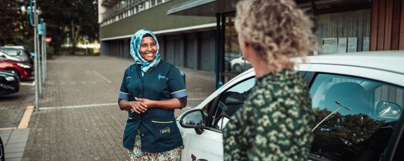 Zorgmedewerker en vrouw staan naast een auto en lachen naar elkaar