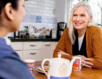 Oudere vrouw zit aan tafel met zorgmedewerker en lacht